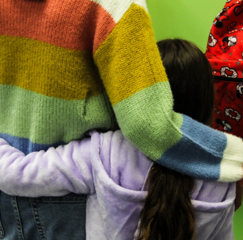 A tween Latina girl in a striped sweater puts her arm around a young Latina girl in a purple sweatshirt