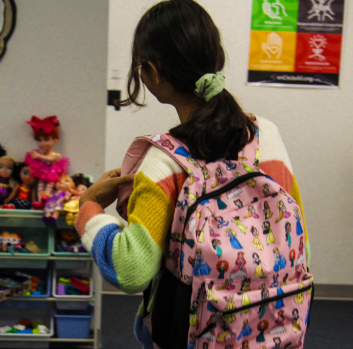 A photo of a pink backpack with Disney princesses on a girl with dark, black hair that is pulled back with a green scrunchie