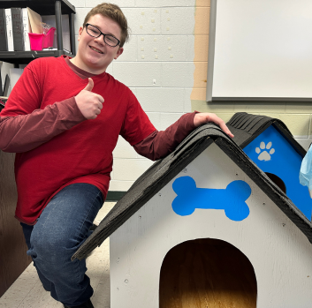 A boy in a red shirt and jeans wearing black glasses gives a thumbs up next to a dog house