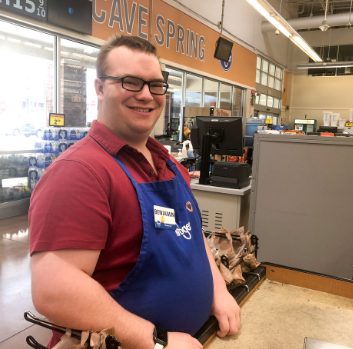 Ben stands in a grocery store in his blue clerk apron and smiles at the camera