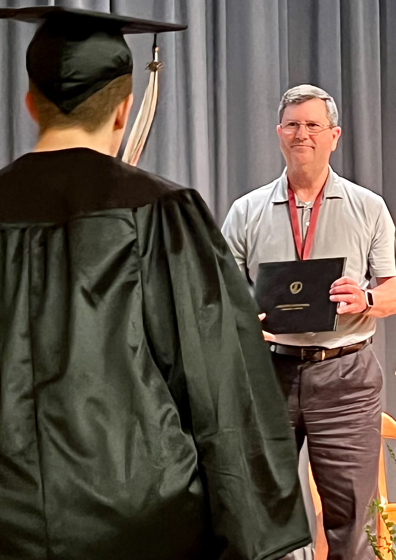 A man holds a diploma to a graduate in a cap and gown whose back is to the camera