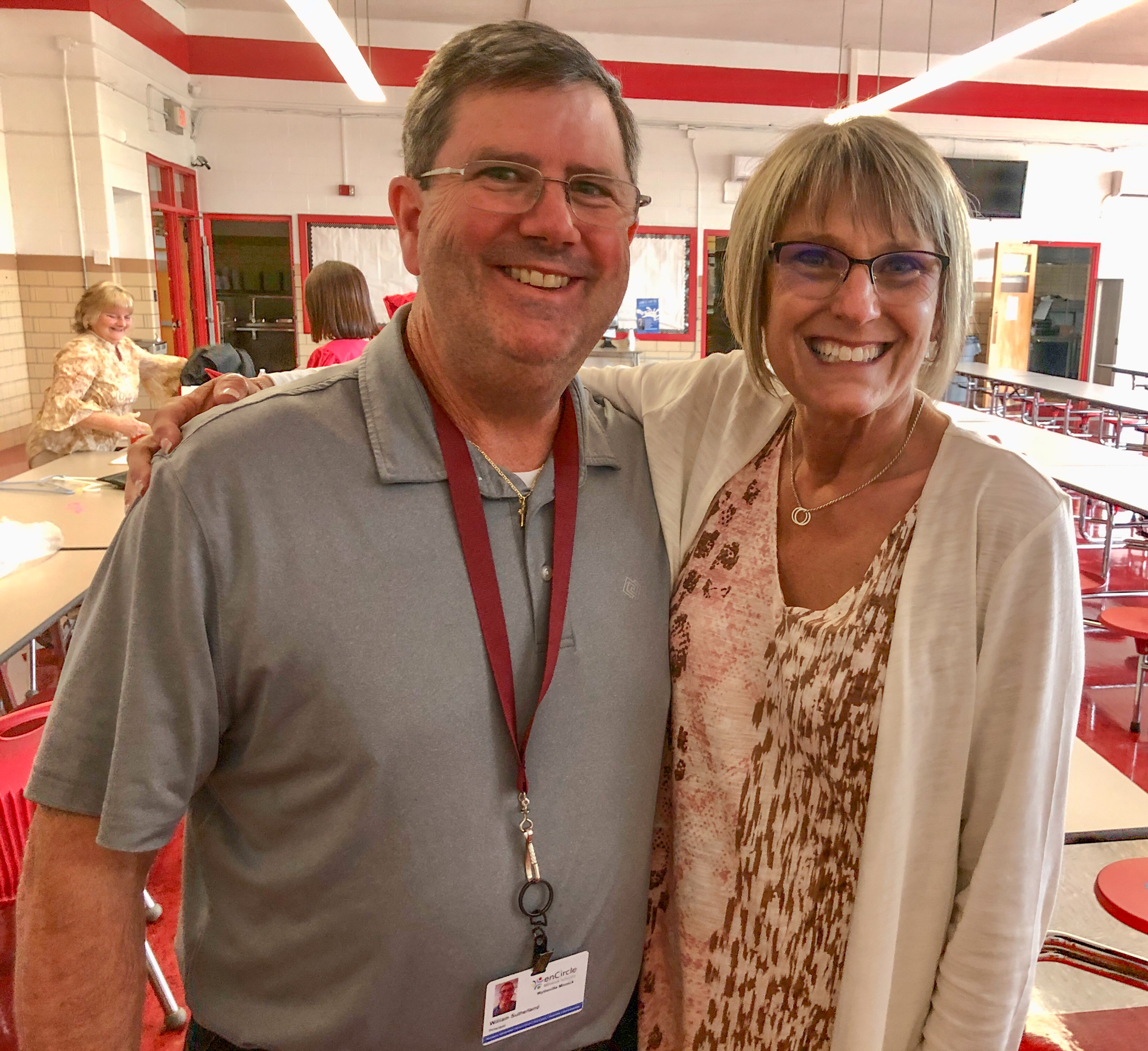 A man and woman stand arm-in-arm in the school cafeteria and smile at the camera w