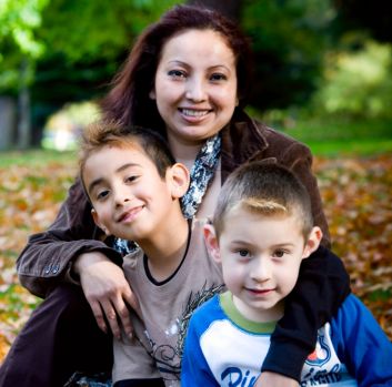 A Latina woman smiles at the camera with her arms around her two young sons