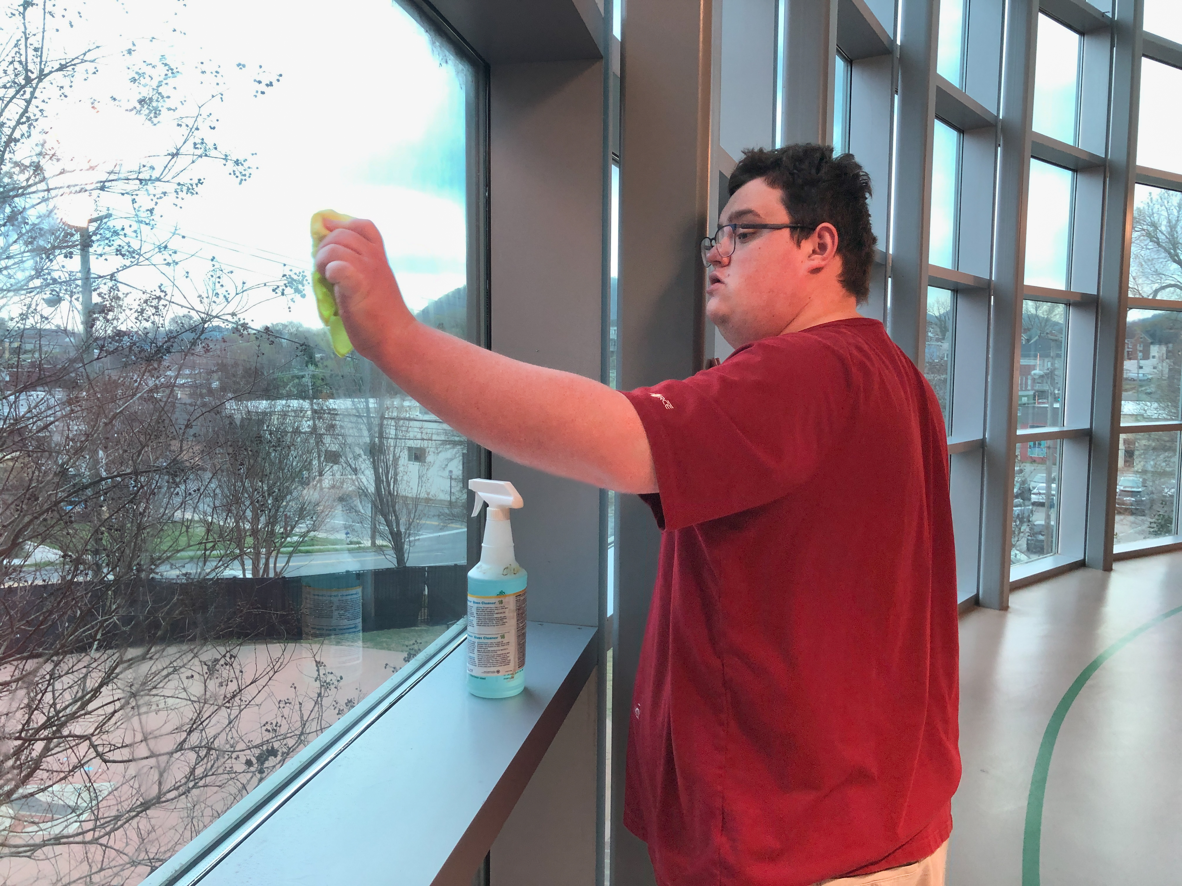 A teenage boy in a red shirt with glasses cleans a window overlooking bare trees