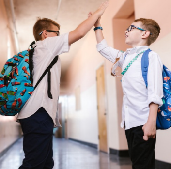 Two young students wearing backpacks high-five in a school hallway