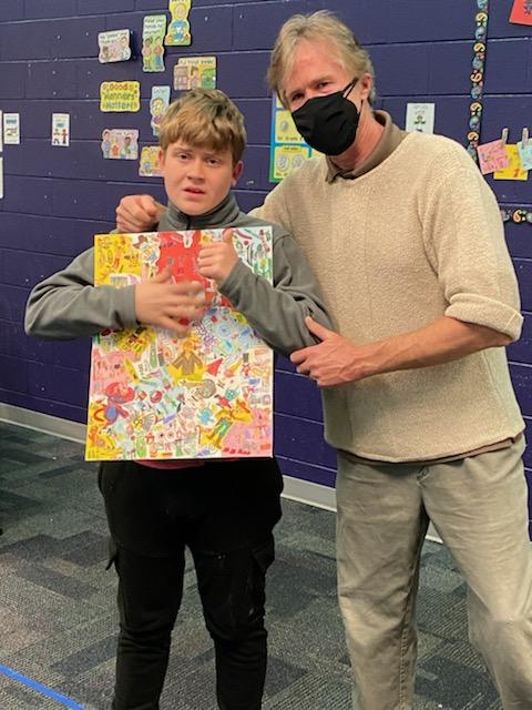 Oliver holds a painting and gives a thumbs up to the camera. His teacher stands with his arm around him.