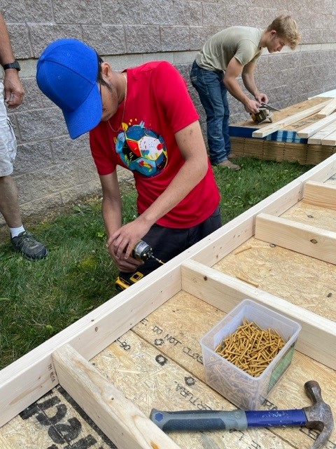 A teen boy wearing a blue baseball camp drills into a two by four piece of wood