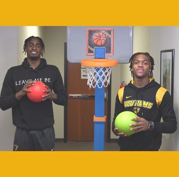 Jalen DeLoach and Jayden Nunn stand smiling in front of a toy basketball hoop inside an office hallway
