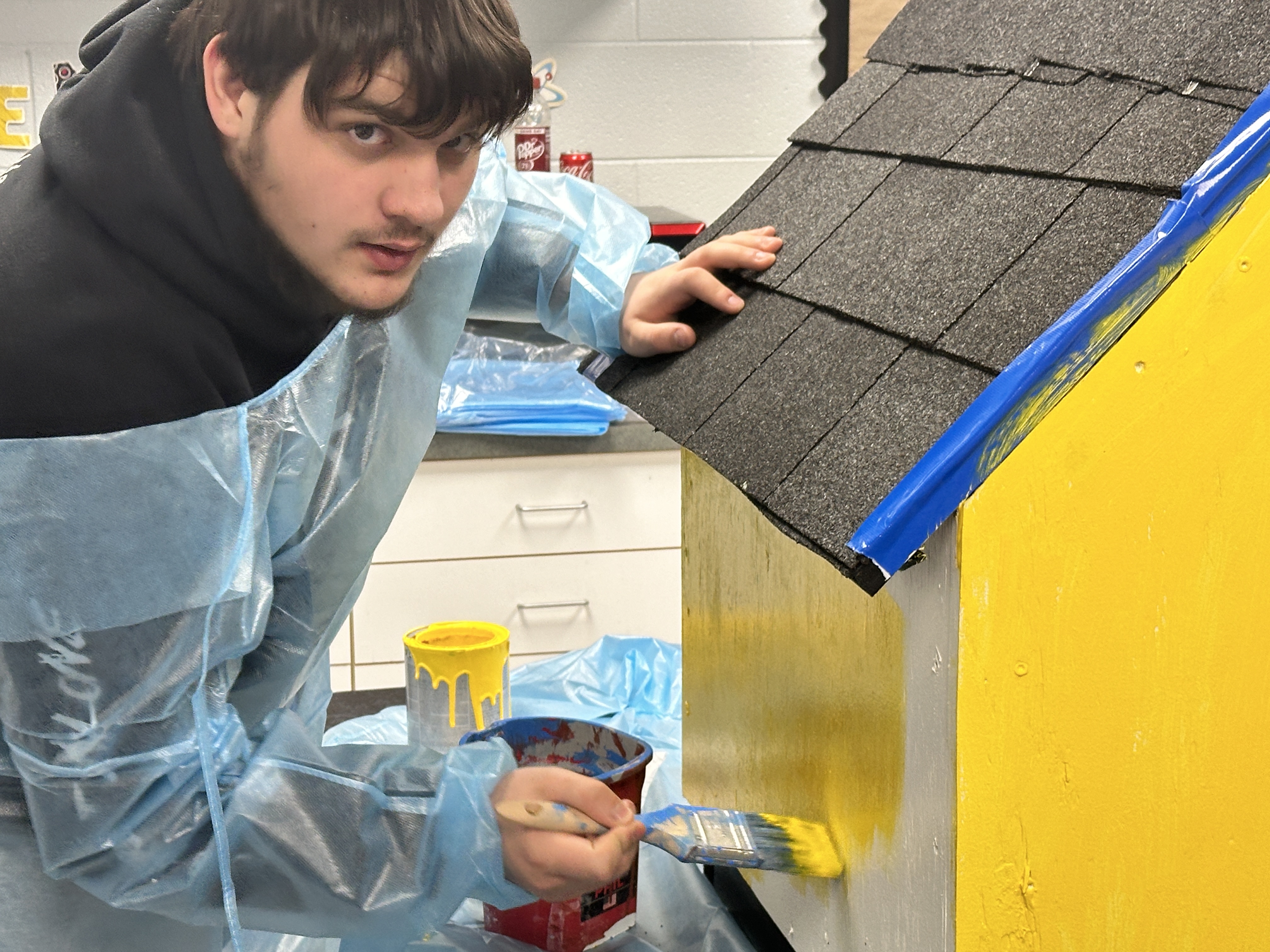 A teen looks at the camera while painting a dog house bright yellow