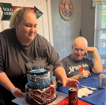 A woman places a two-tier cake on a table in front of an older man wearing a Spiderman shirt