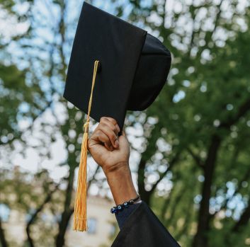 A hand holds up a graduation hat in front of a background of trees