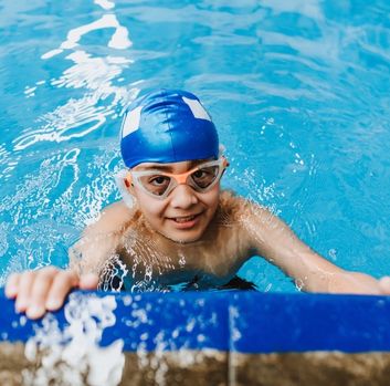 A young Hispanic boy wearing goggles and blue swim cap holds onto the side of a pool and looks at the camera