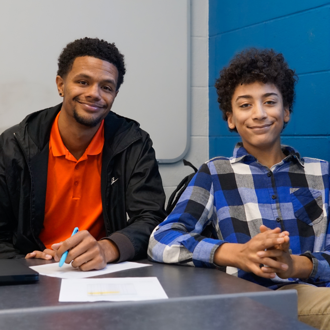 A black teacher and black teenage student sit together at a table with worksheets and smile at the camera