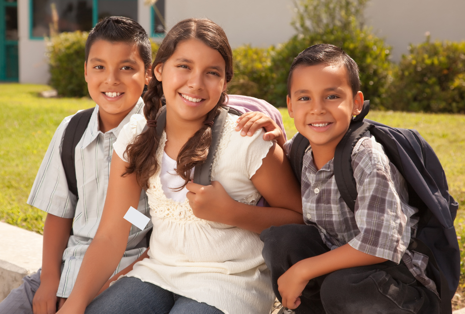 Two Latino boys sit on either side of a Latina teen wearing backpacks outside