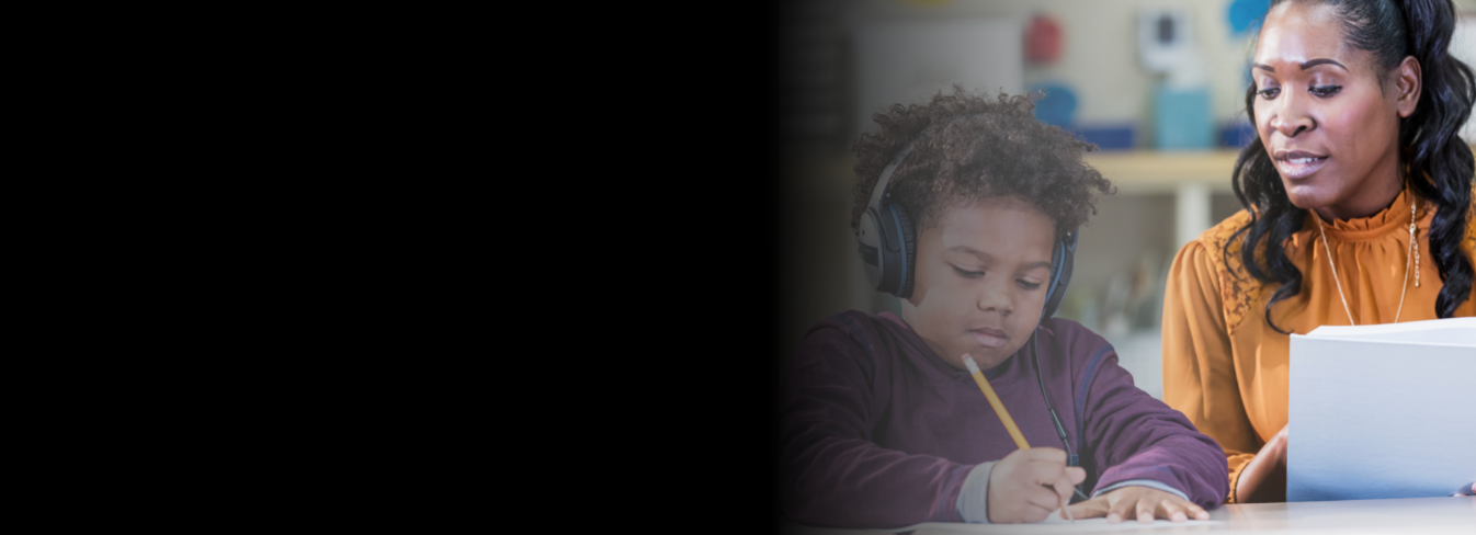 An African American teacher looks over the shoulder of an young, African American student who is wearing headphones and writing with a pencil