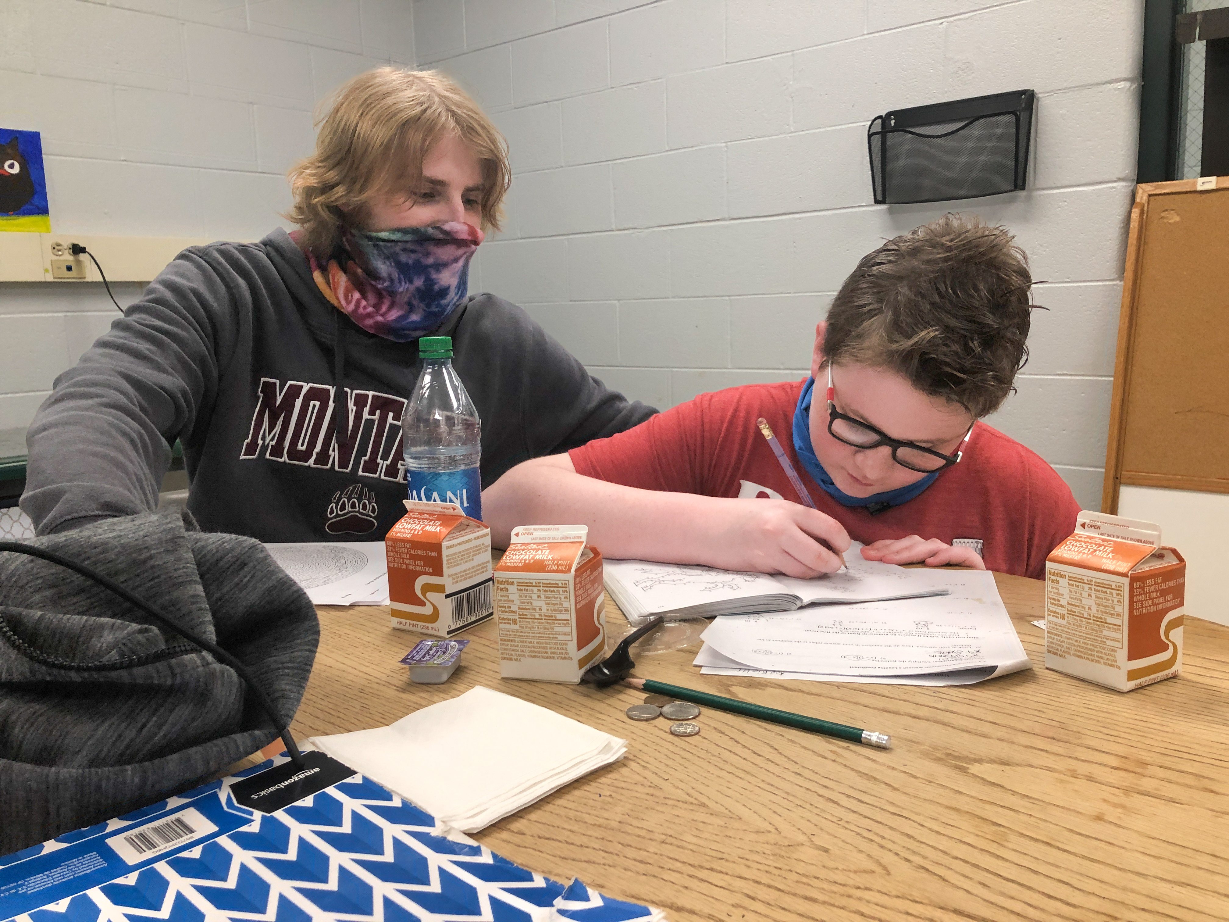 A teacher wearing a mask leans over the shoulder of a student who is writing in a notebook