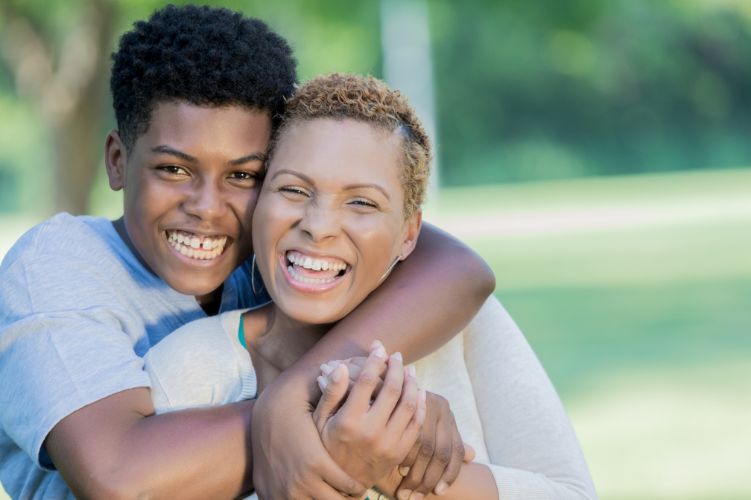 An African American teen smiles and hugs an African American woman