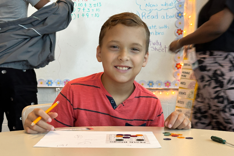 A young white boy with a pencil in his hand sits in a classroom and smiles at the camera