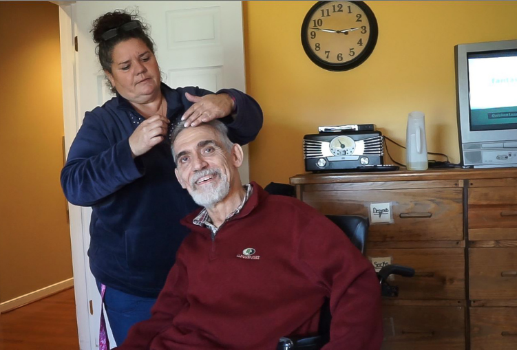 A man in a wheelchair smiles as a woman combs his hair. They are in home setting.