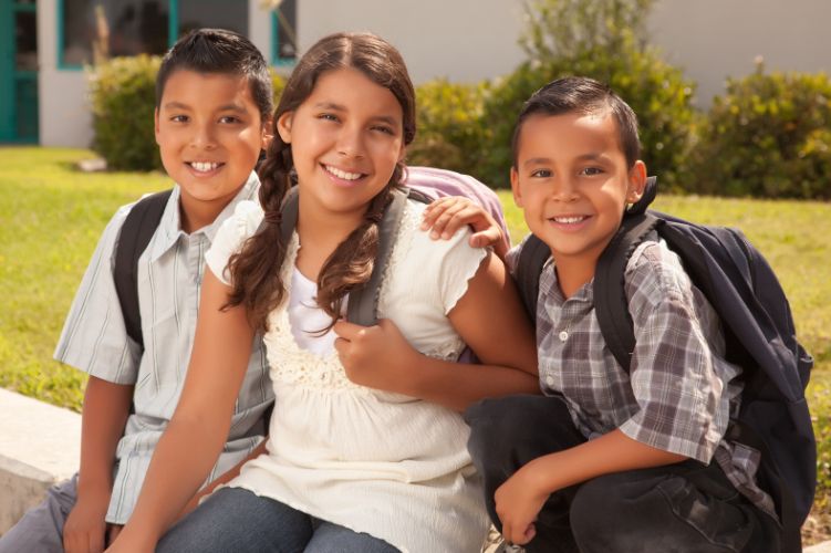 Two Latino boys and a Latina girl sit outside wearing backpacks and smiling at the camera