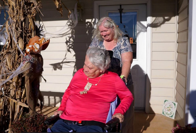 A woman pushes another woman in a wheel chair down a front porch ramp