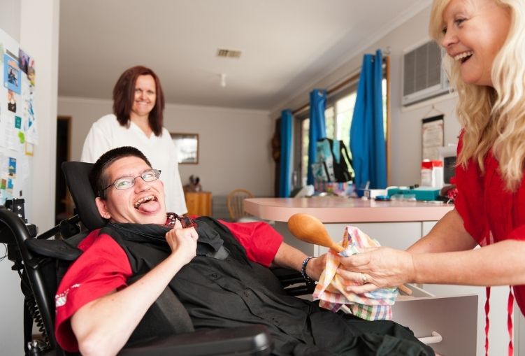 A smiling woman in a kitchen hands a corn dog to a man in a wheel chair with a developmental disability. Another woman stands in the distance and smiles at them.
