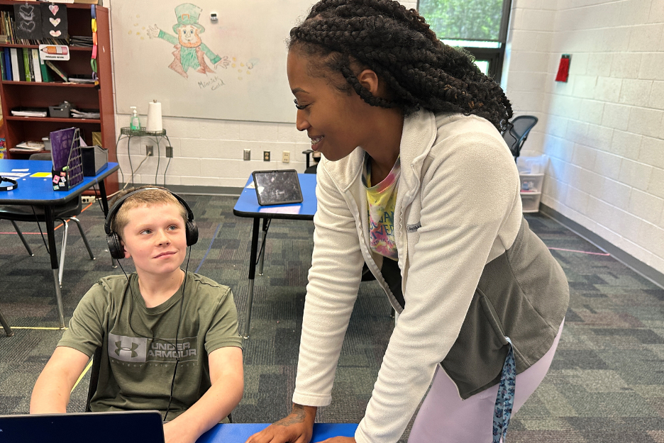 A student sits at a desk with a laptop and smiles up at his teacher