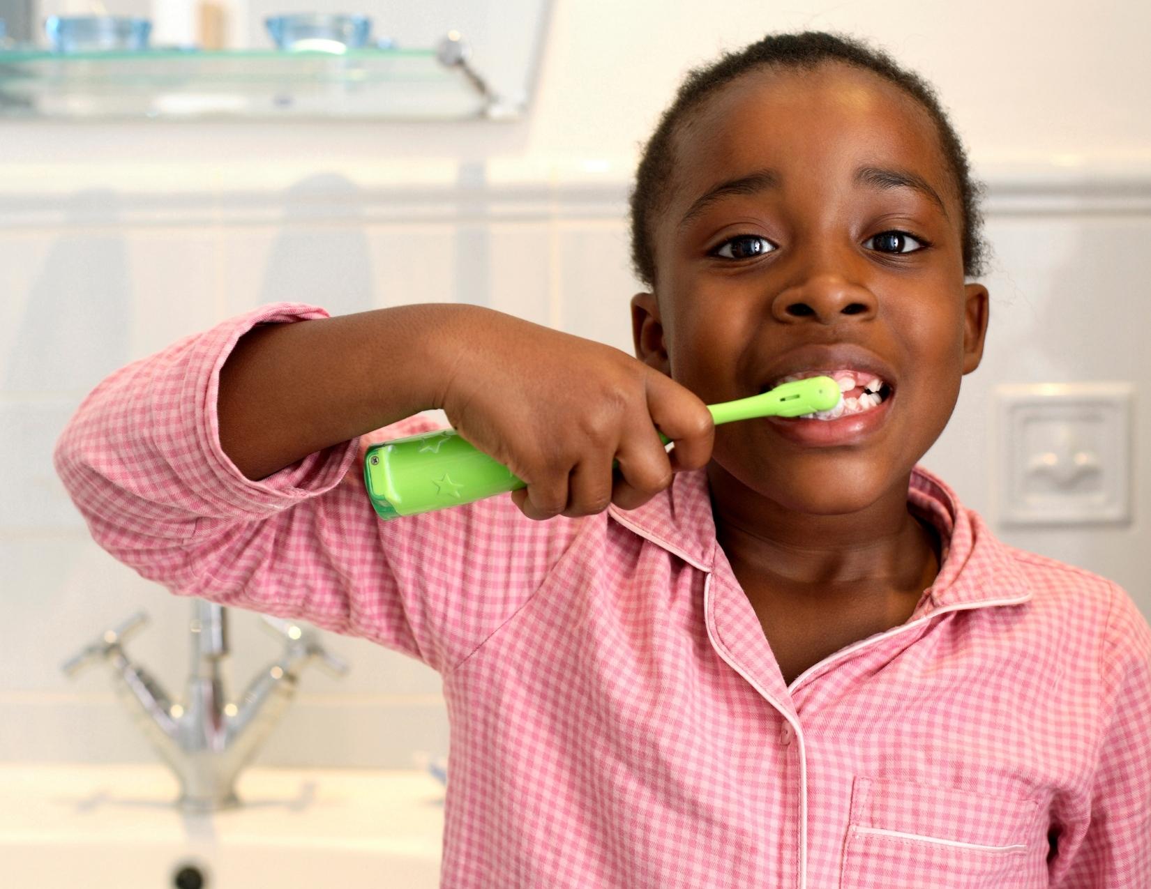 A black child brushes their teeth in the bathroom with a green toothbrush while wearing light red checked button-up pajamas