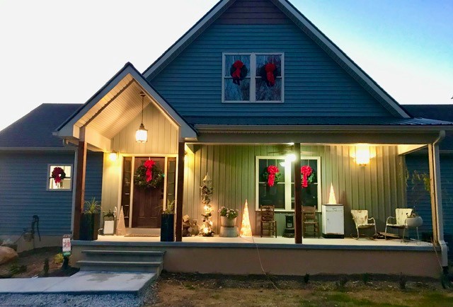 Farm style house with front porch at dusk. Porch lights are on and green wreaths with red ribbons hang on the windwos.
